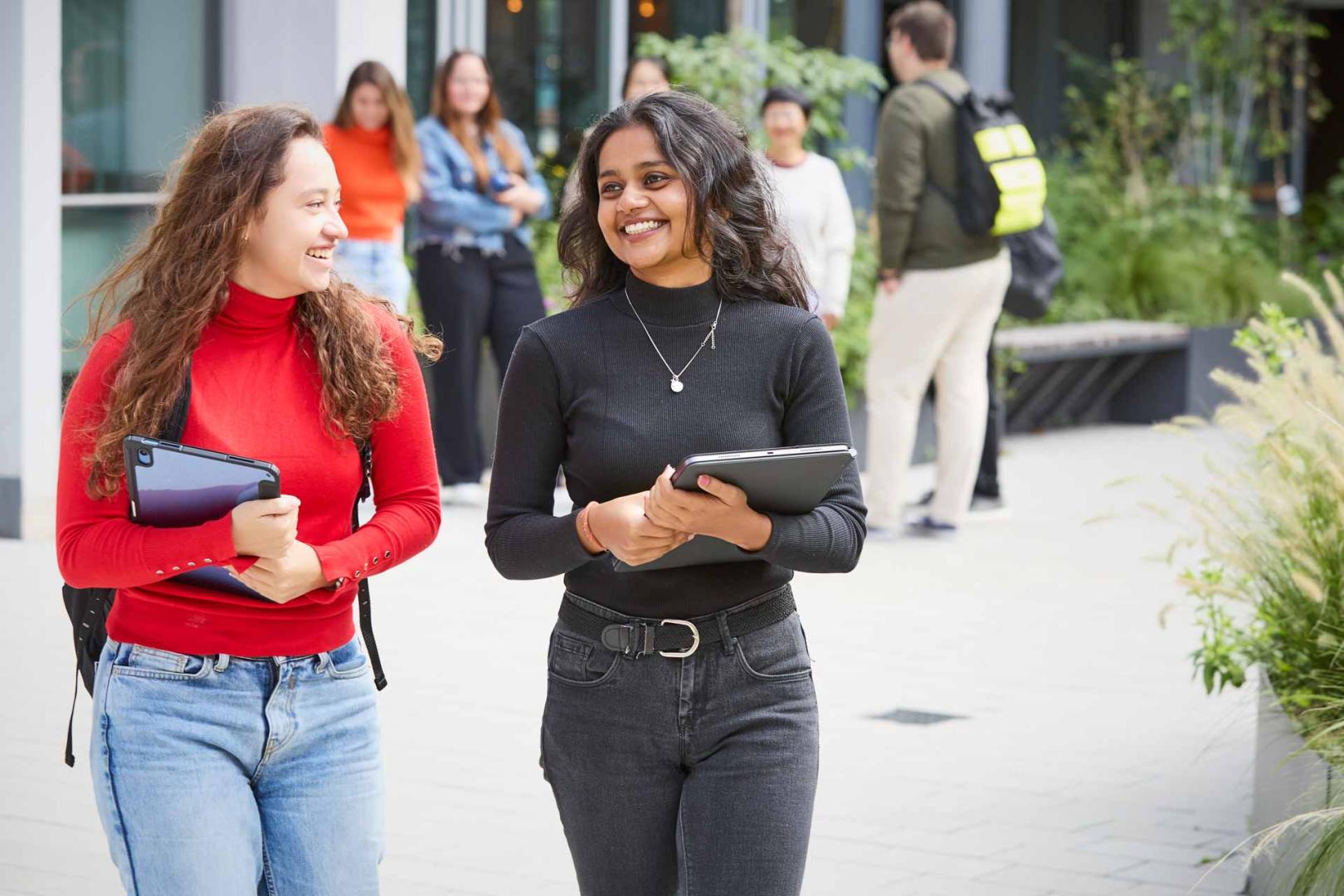 Zwei Studentinnen gehen lachend über den Campus, umgeben von anderen Studierenden. Die Szene strahlt eine freundliche und lebendige Studienatmosphäre aus.