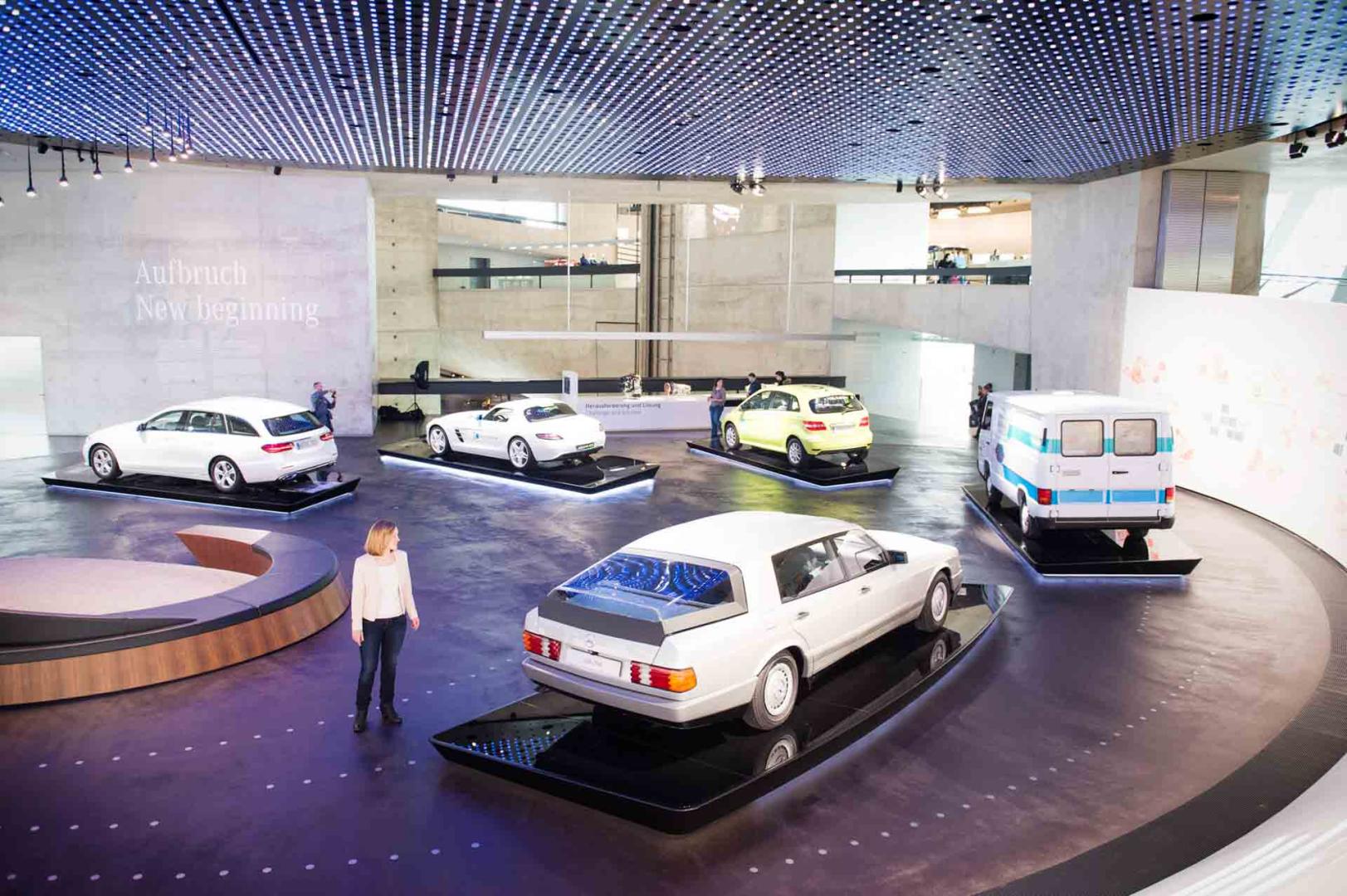 Frau steht im Mercedes-Benz Museum, umgeben von historischen Autos. Die Perspektive zeigt die Frau aus der Vogelperspektive, was ihre kleine Größe im Verhältnis zu den beeindruckenden Fahrzeugen betont.