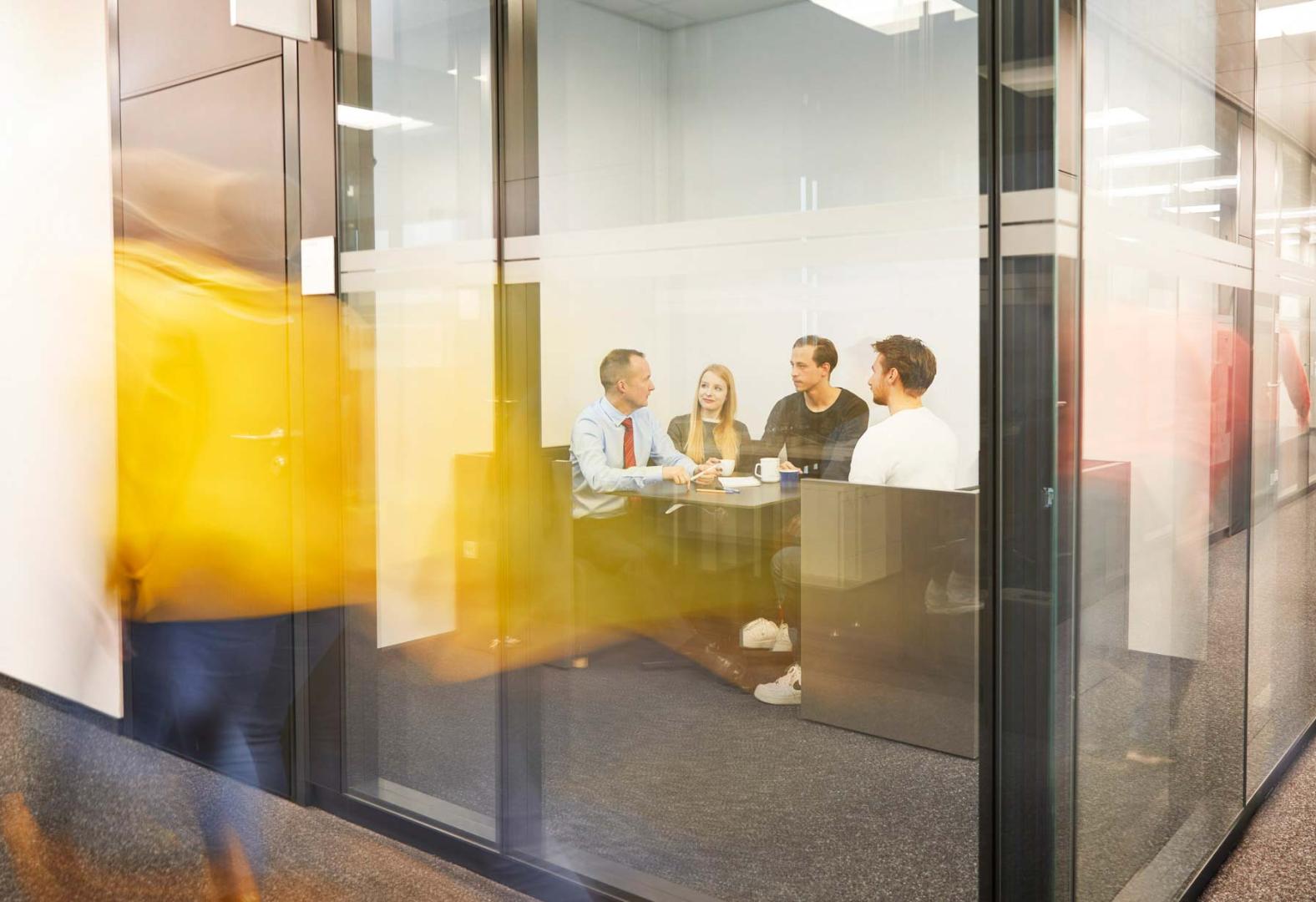 Moderne Besprechungssituation: Mehrere Personen sitzen in einem verglasten Meetingraum und diskutieren. Durch die transparente Glaswand ist eine Person im Außenbereich zu sehen, die vorbeiläuft. Das Bild vermittelt eine dynamische Arbeitsatmosphäre in einem modernen Büro.